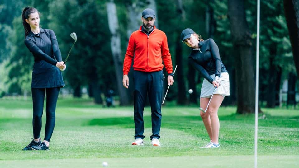Two woman golfers are seen practicing how to chip a golf ball with the help of a golf coach.