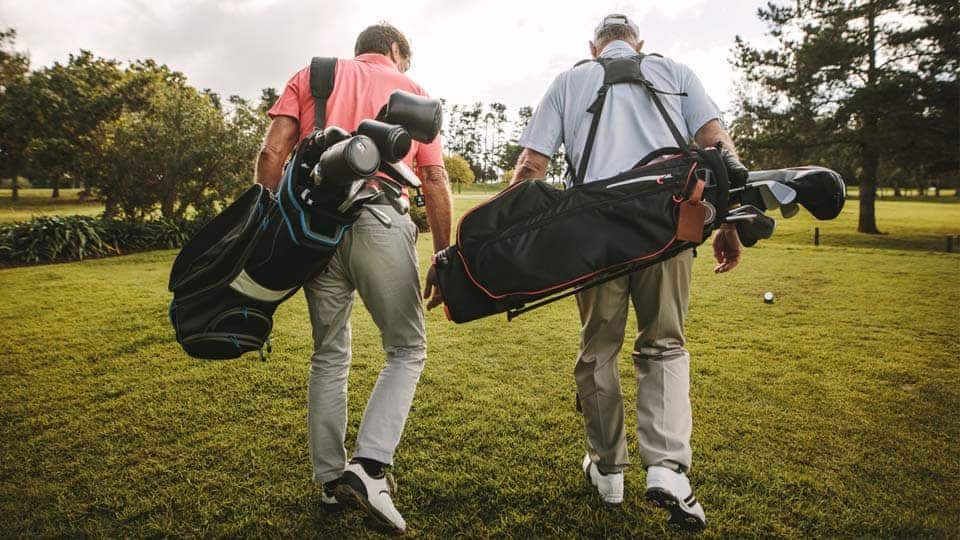 Two golfers walking towards the tee with the golf bag filled with clubs on their back.