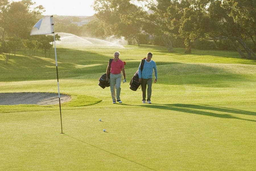 Two golfers are walking in the golf course with their golf bags near a golf hole