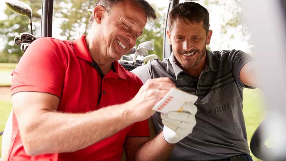 Two golfers sitting in a golf cart with their clubs and calculating their golf handicap.