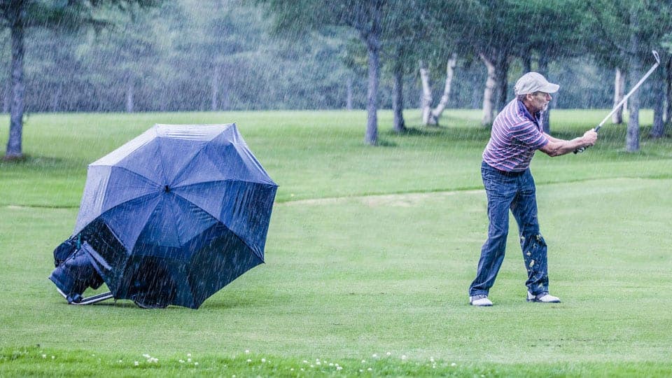 A golfer chipping shot in the rain with a broad umbrella covering the golf club bag placed on the course.
